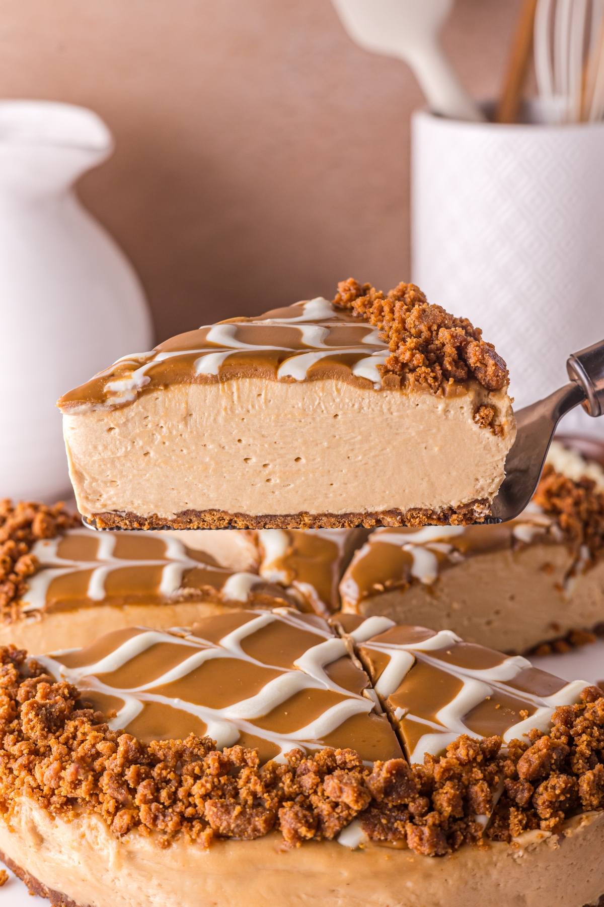 A slice of creamy caramel no bake biscoff  cheesecake with cookie crust and white drizzle is lifted from the whole cake.