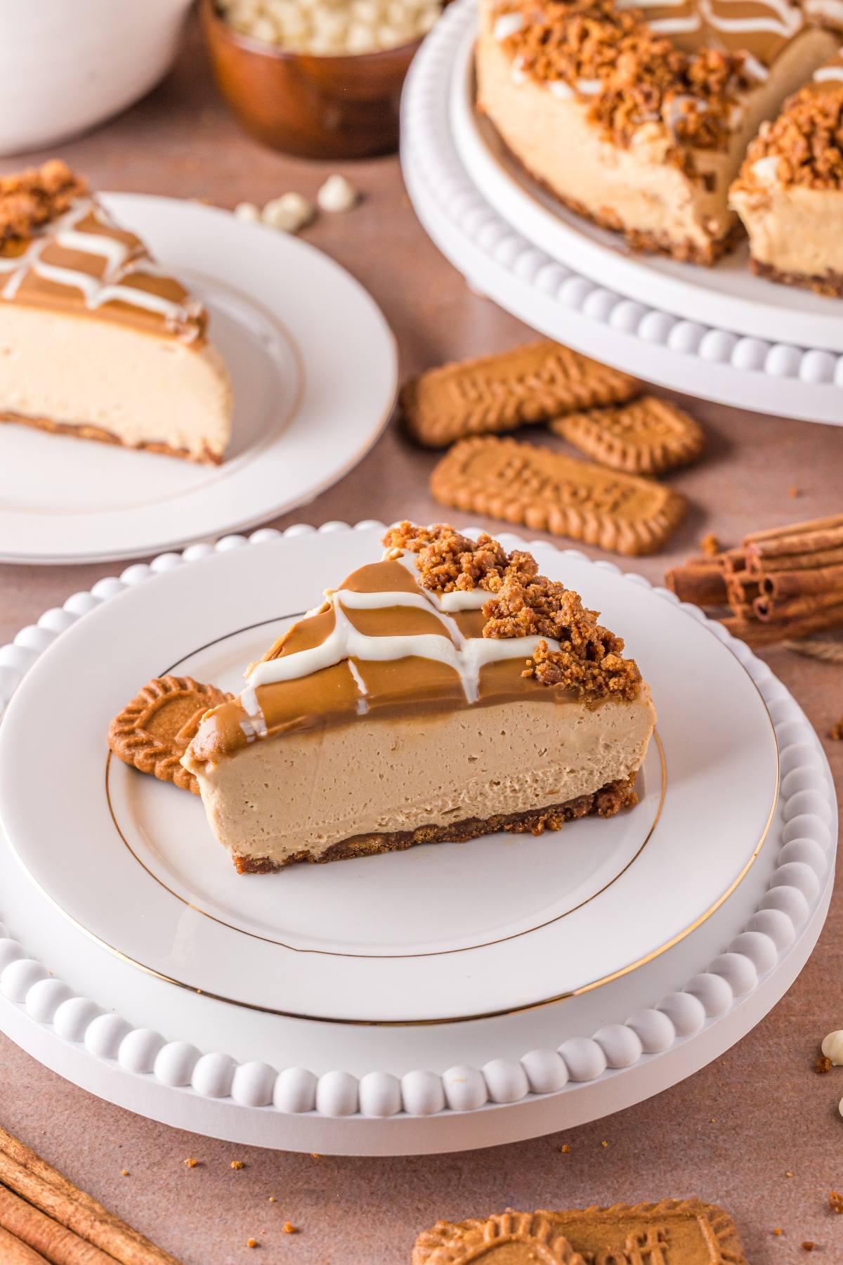 A slice of no bake biscoff  cheesecake topped with caramel, crumbs, and white drizzle on a plate with a cookie.