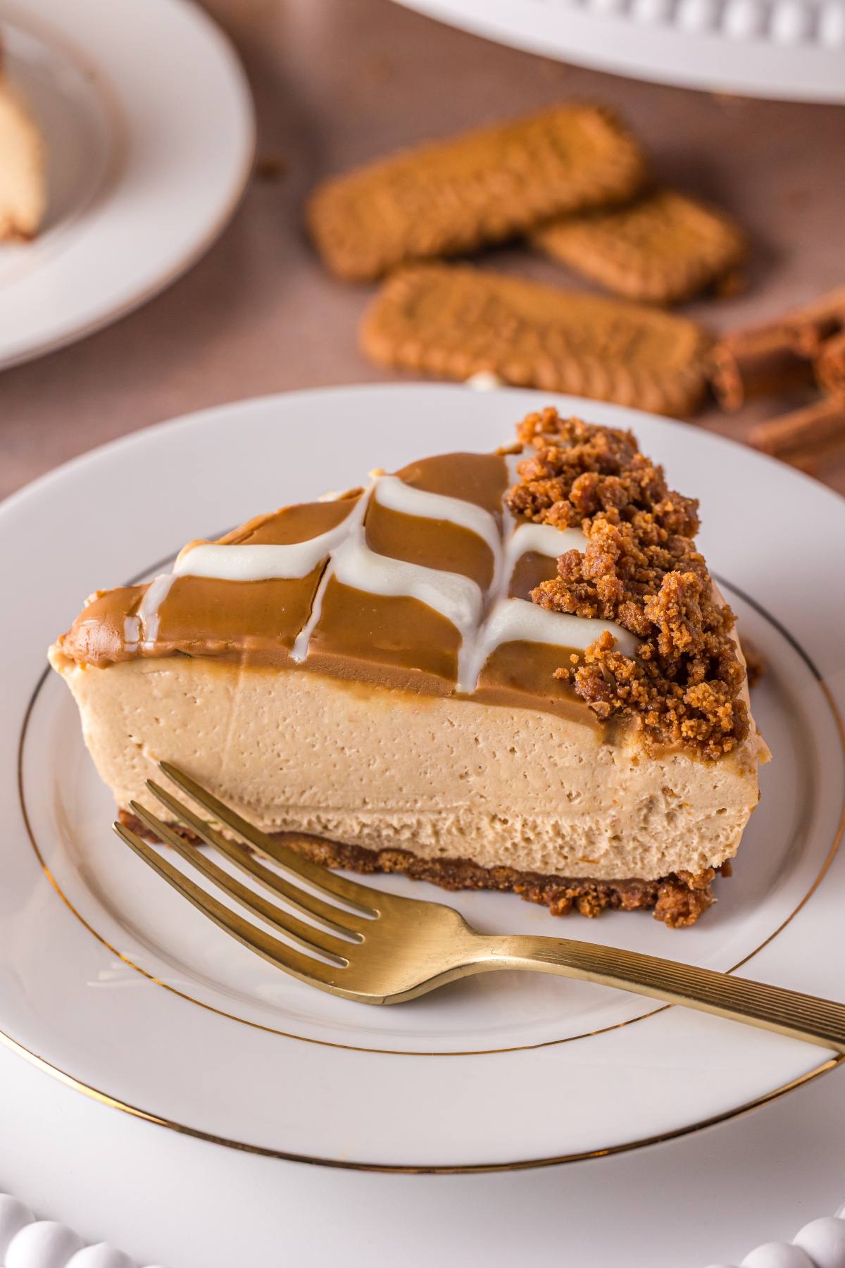 A slice of no bake biscoff  cheesecake with caramel drizzle and crumbled cookies on a white plate with a gold fork.