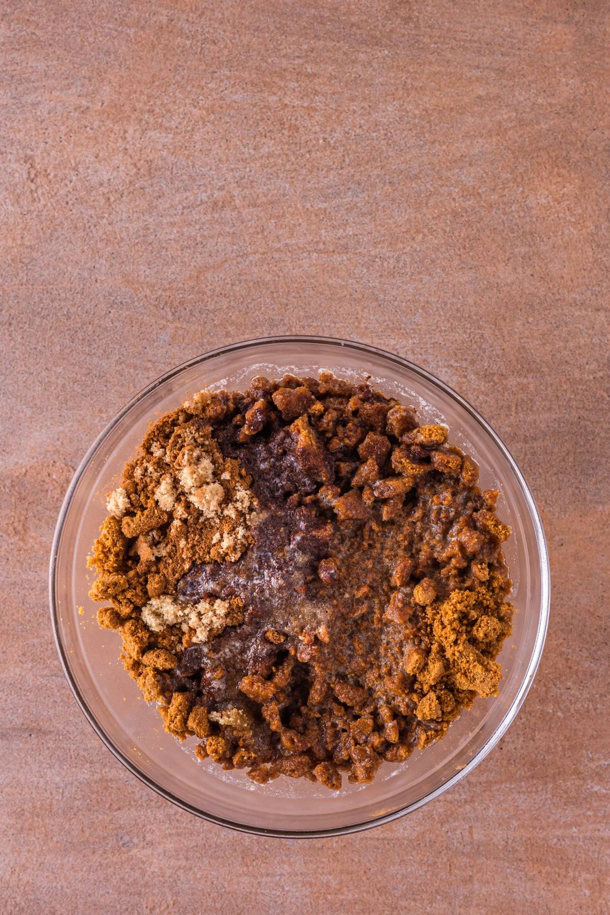 A glass bowl with brown sugar, spices, and liquid ingredients on a brown surface, viewed from above.