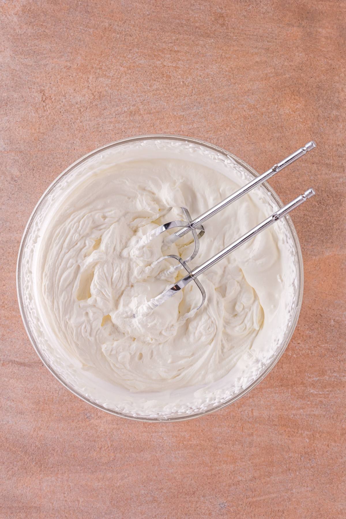 A glass bowl of whipped cream with metal beaters on a brown countertop, viewed from above.