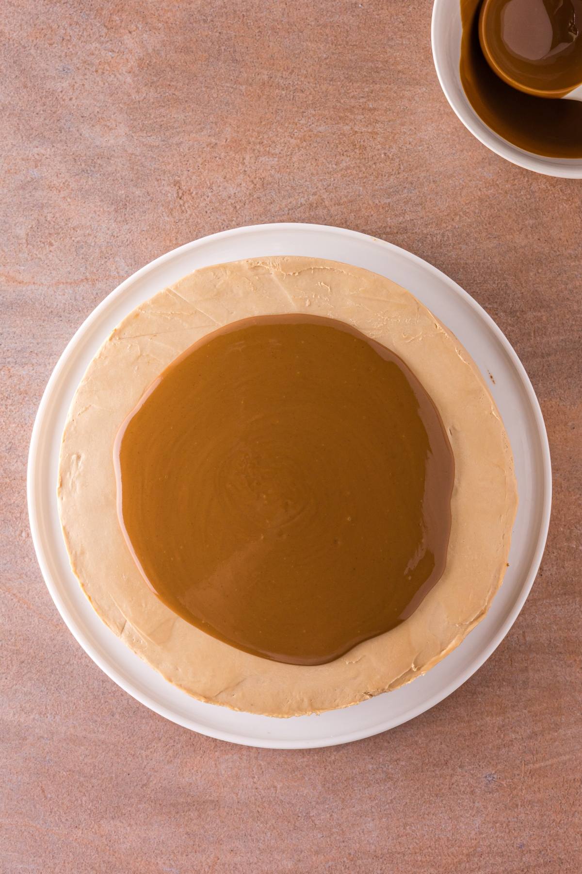A round cake with caramel glaze on top, placed on a white plate, with a bowl of caramel sauce nearby.