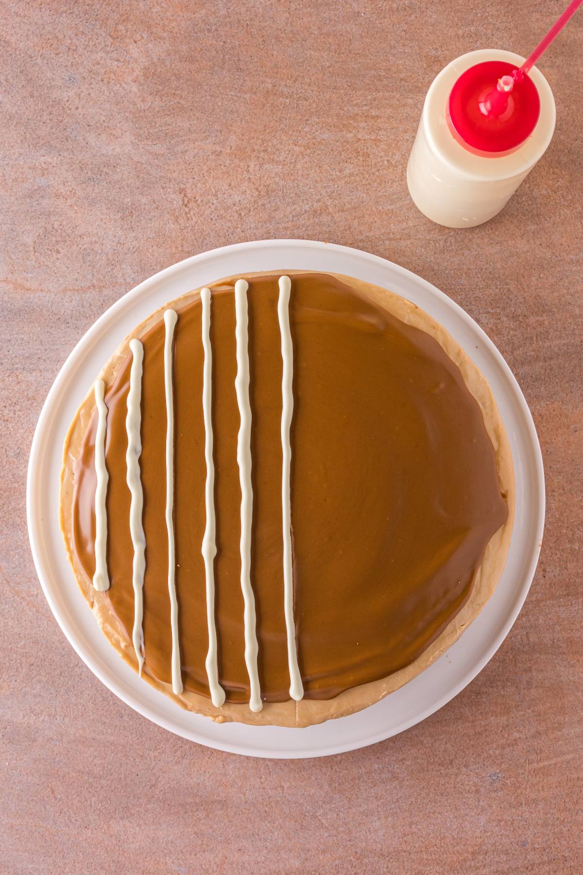 Round cake with caramel glaze, decorated with white icing lines on one side, next to a bottle of white sauce.