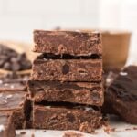 A stack of thick, homemade chocolate fudge pieces with sea salt on top, surrounded by more fudge and no bake brownie bars studded with chocolate chips.