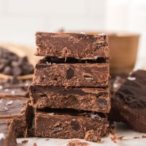 A stack of thick, homemade chocolate fudge pieces with sea salt on top, surrounded by more fudge and no bake brownie bars studded with chocolate chips.