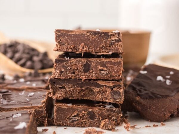 A stack of thick, homemade chocolate fudge pieces with sea salt on top, surrounded by more fudge and no bake brownie bars studded with chocolate chips.