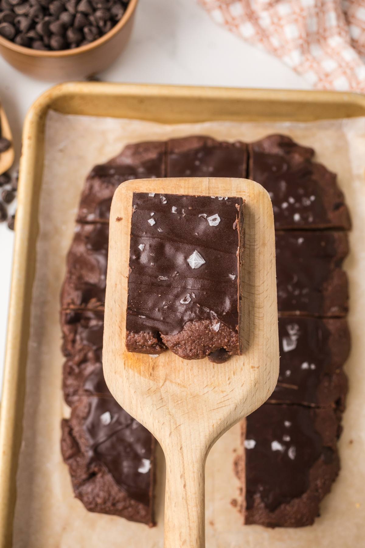 A wooden spatula lifts a chocolate-covered brownie with flaky sea salt from a tray of brownies.
