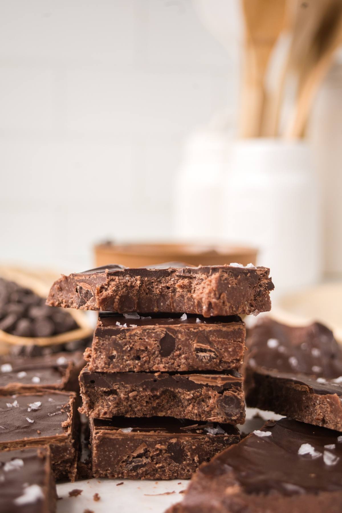 A stack of chocolate no bake brownie bars topped with sea salt, with more fudge and chocolate chips in the background.