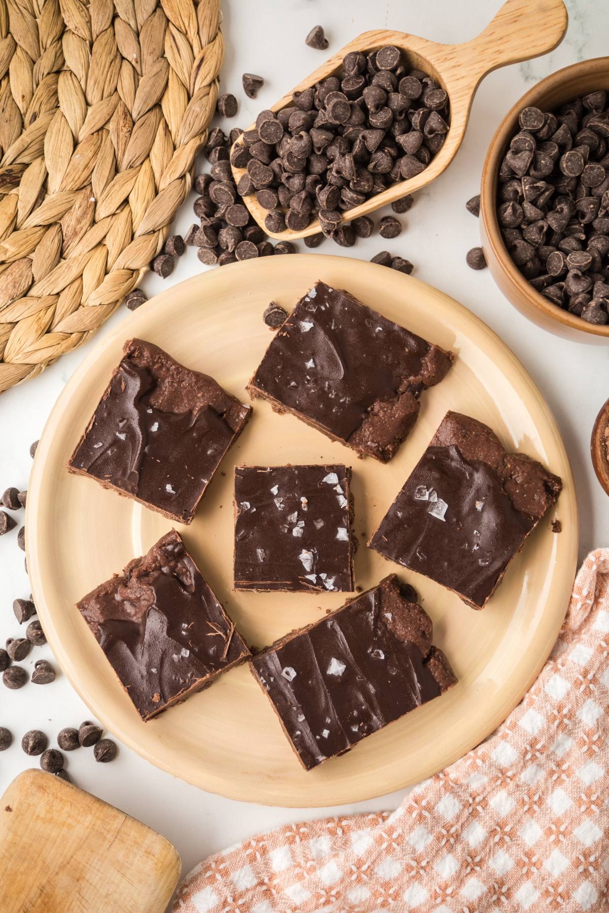 Six chocolate no bake brownie bars  on a plate, surrounded by chocolate chips and kitchen items on a table.