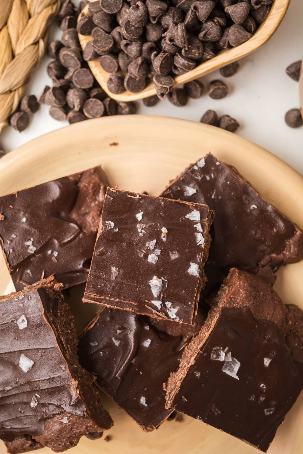 Five chocolate no bake brownies bars topped with flaky sea salt on a wooden plate, with chocolate chips nearby.