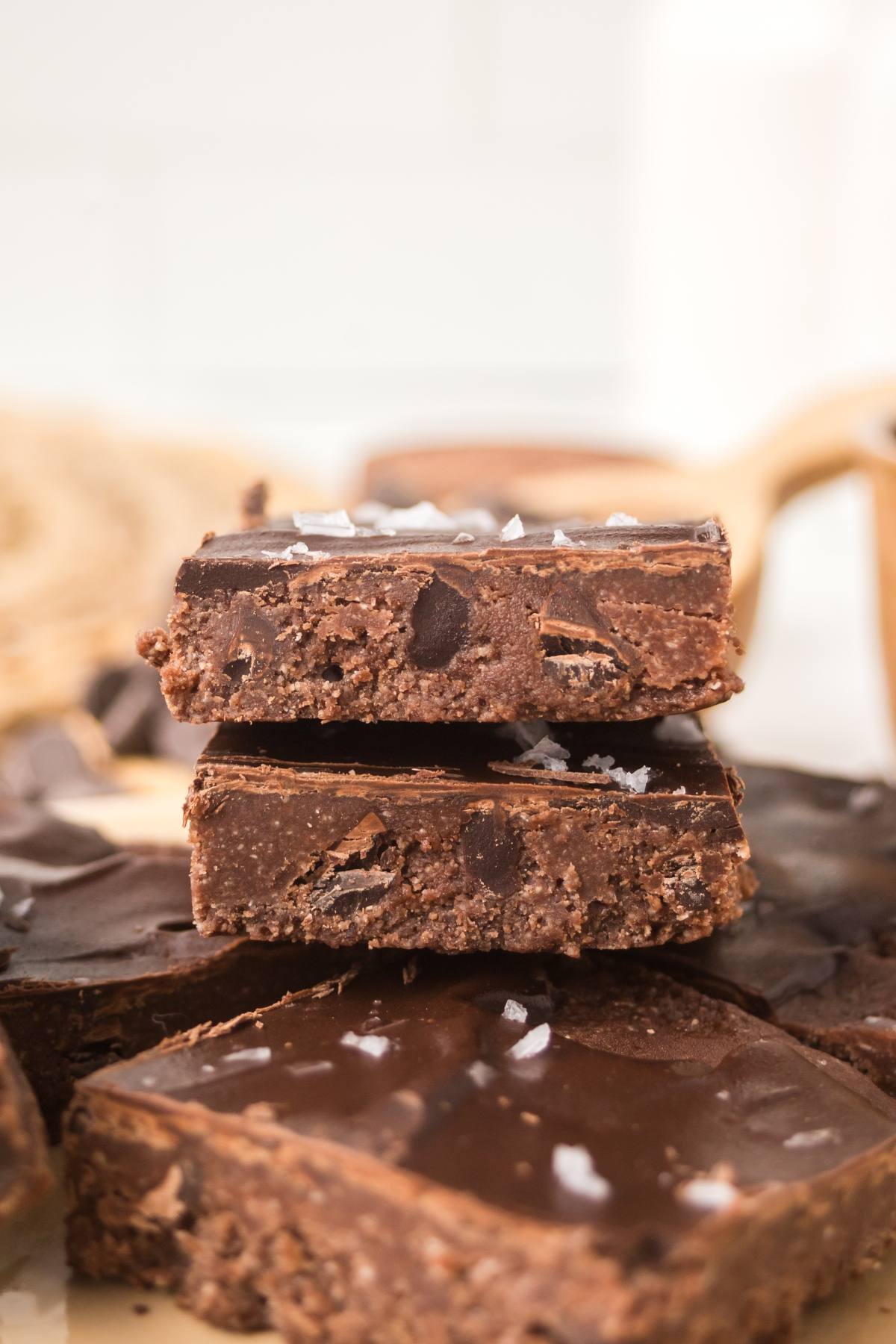 Three thick, chocolate fudge bars stacked, topped with flaky sea salt, with more bars in the background.