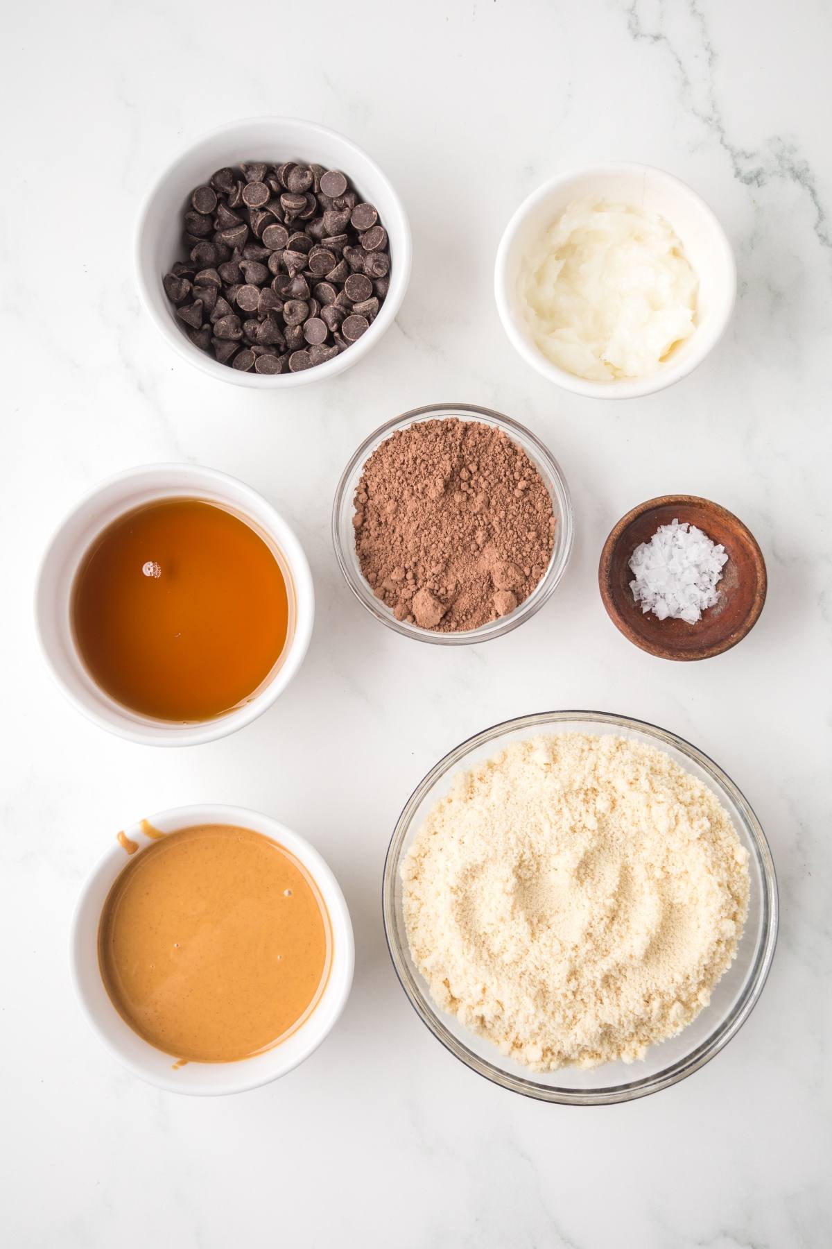 Seven bowls with chocolate chips, almond flour, cocoa, peanut butter, maple syrup, coconut oil, and salt on a counter.