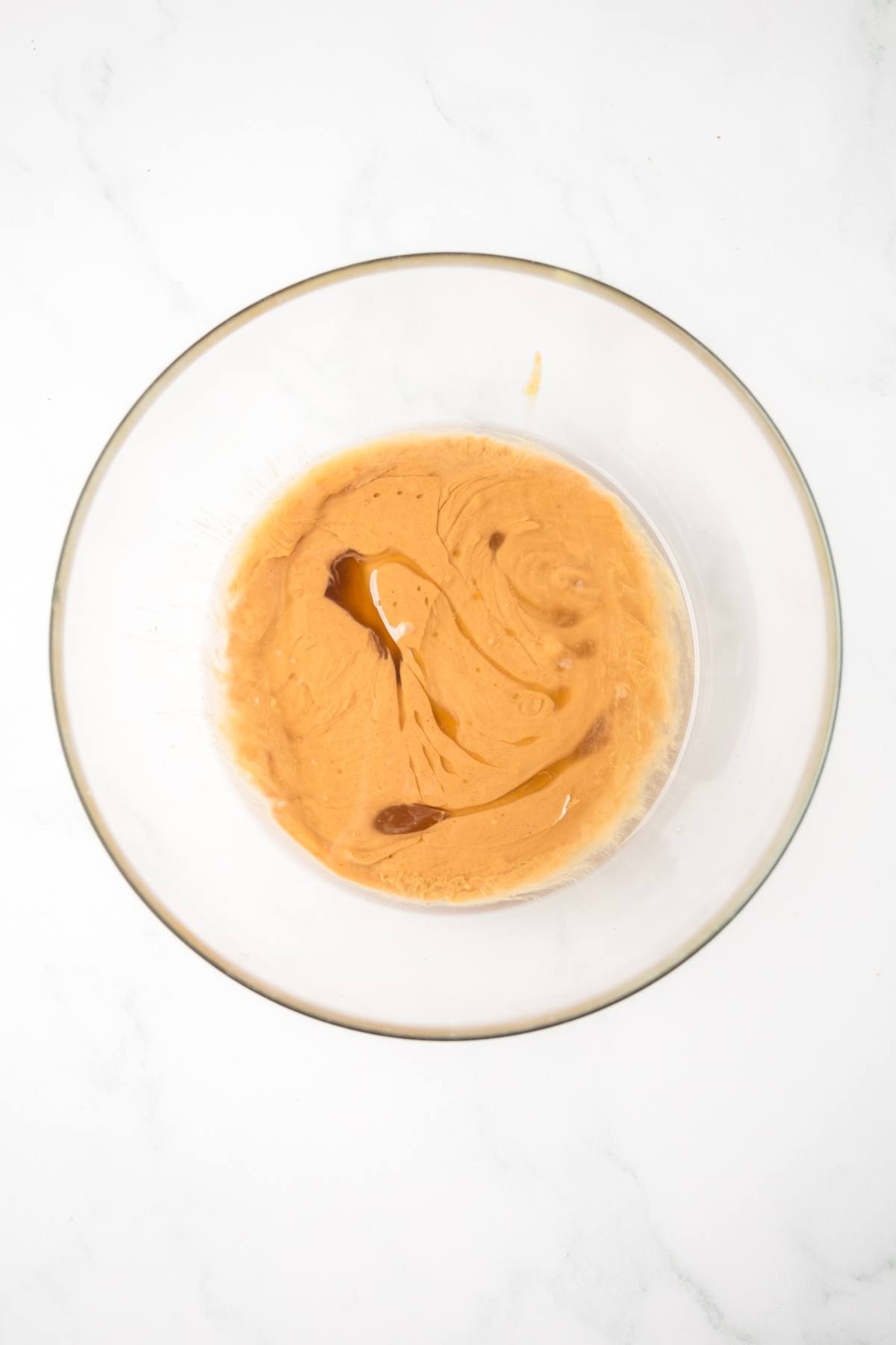 Glass bowl with light brown batter and a swirl of vanilla extract on a white surface.