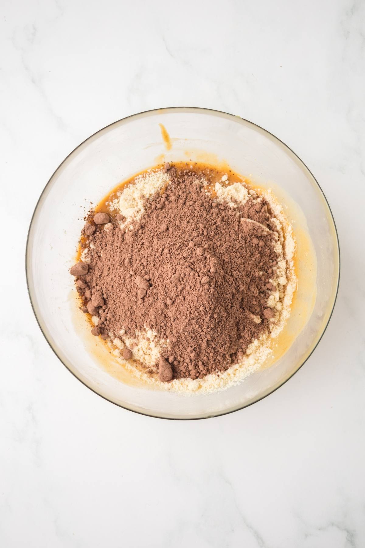 Glass bowl with cocoa powder, flour, and wet ingredients on a white marble surface, ready to be mixed.