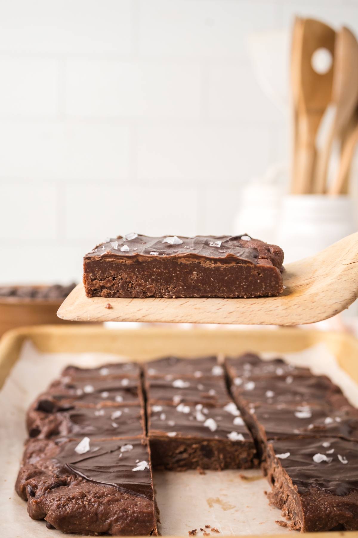 A wooden spatula lifts a chocolate  no bake brownie bars topped with sea salt from a tray of sliced brownies.