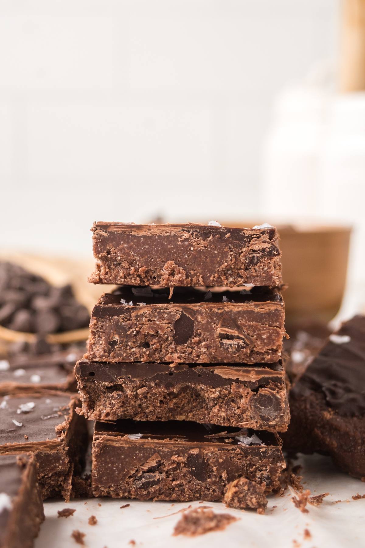 A stack of four chocolate no bake brownie bars squares with sea salt on top, surrounded by more fudge pieces.