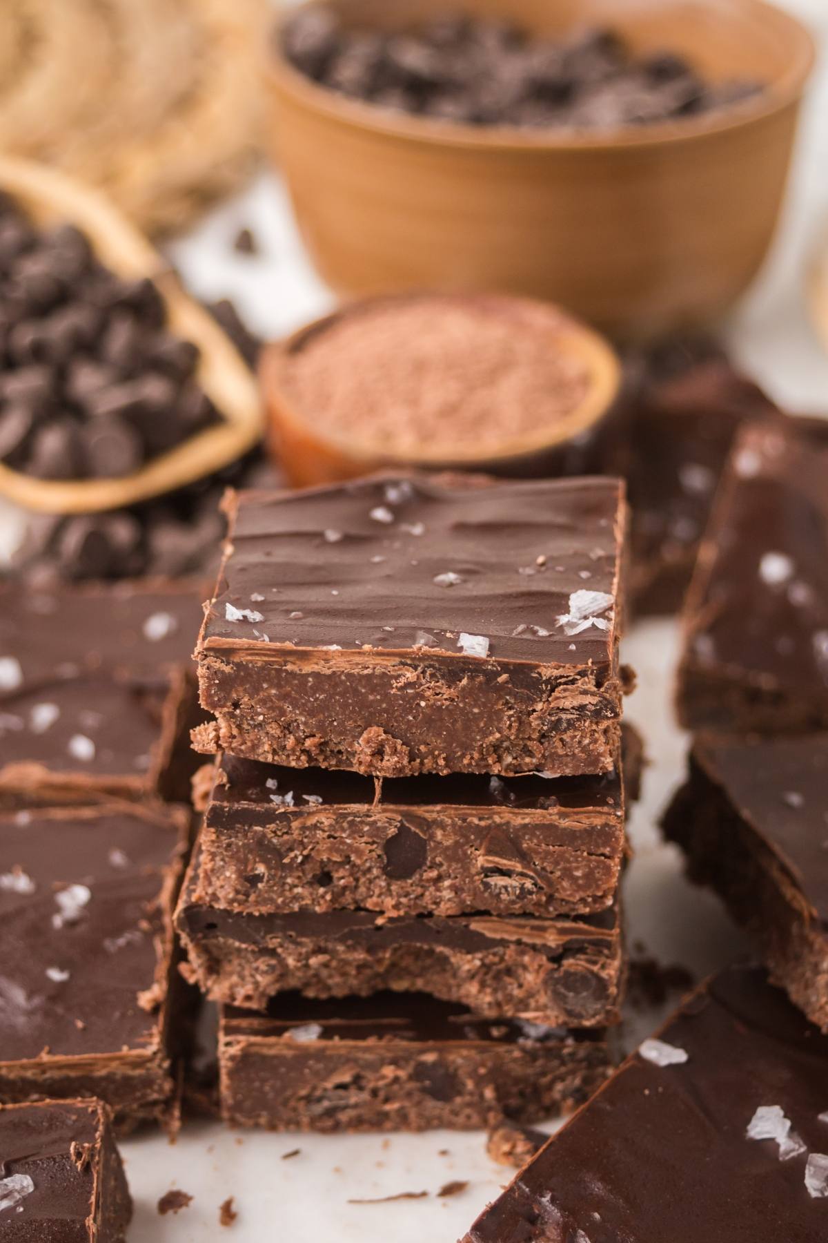 Stacked chocolate no bake brownie bars  topped with sea salt, surrounded by chocolate chips and cocoa powder.
