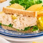 Pepperoncini chicken salad sandwich with lettuce on whole wheat bread, served with potato chips and a pepper on a blue plate.