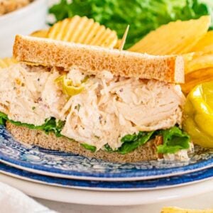 Pepperoncini chicken salad sandwich with lettuce on whole wheat bread, served with potato chips and a pepper on a blue plate.