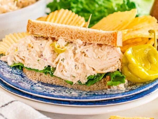 Pepperoncini chicken salad sandwich with lettuce on whole wheat bread, served with potato chips and a pepper on a blue plate.