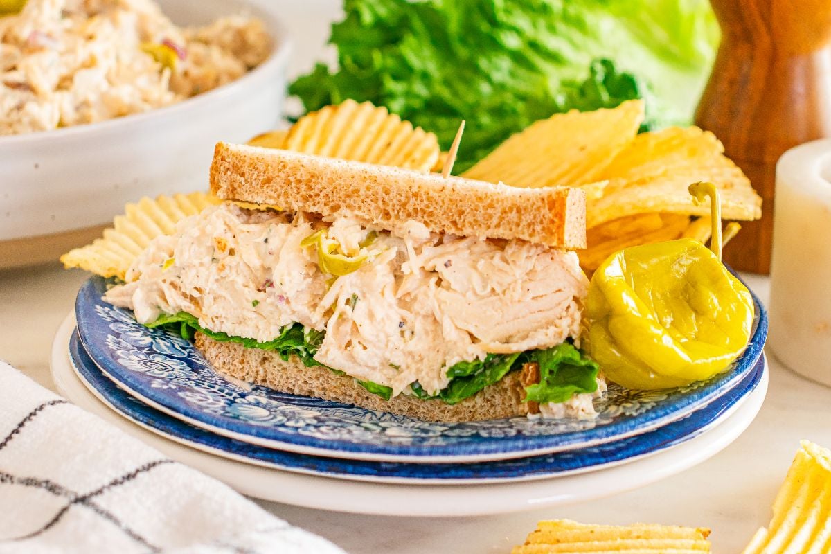 Pepperoncini chicken salad sandwich on wheat bread with lettuce, served alongside potato chips and a crisp pepperoncini on a blue plate.