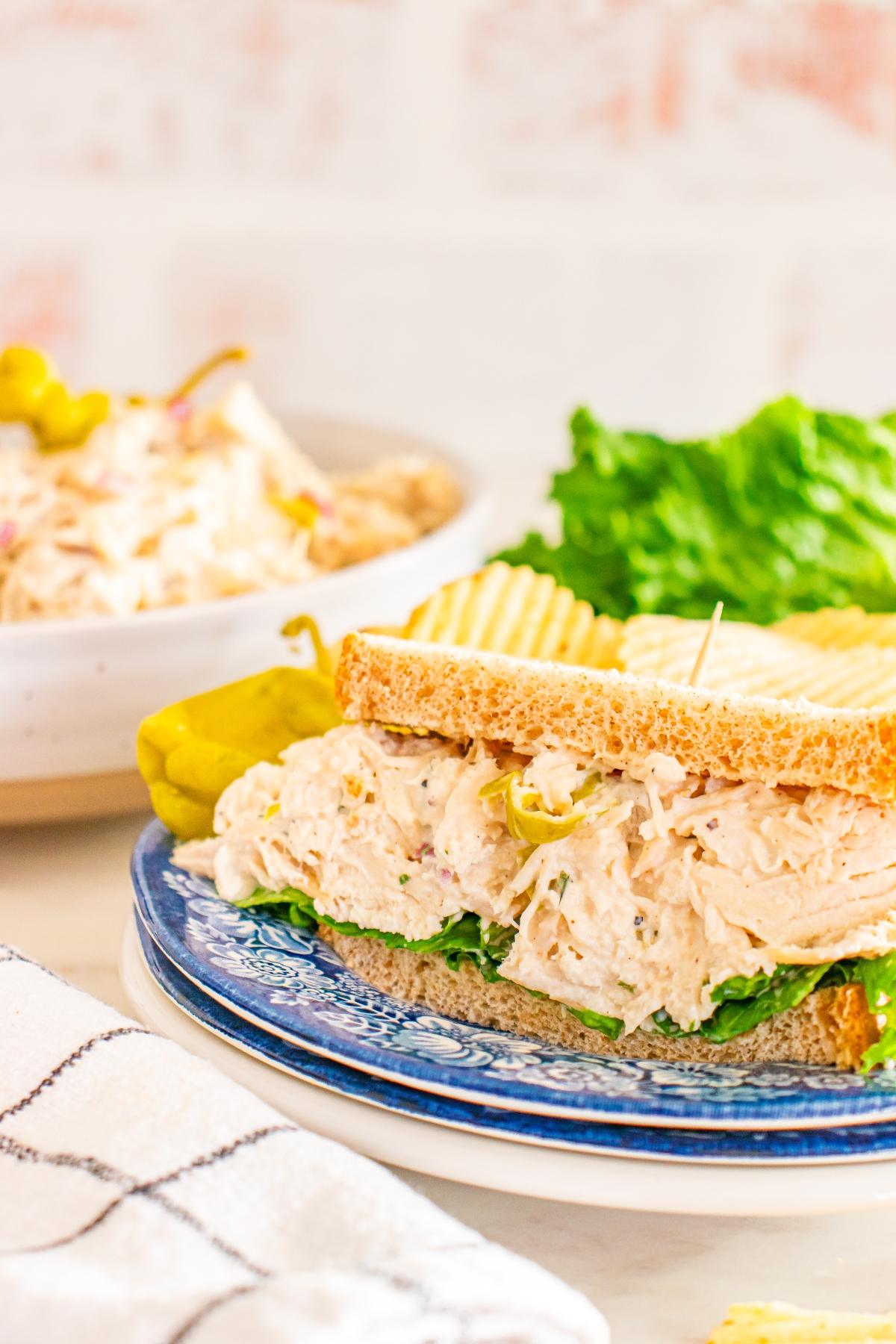 Pepperoncini Chicken salad sandwich on wheat bread with lettuce, served with potato chips and a pickle on a blue plate.
