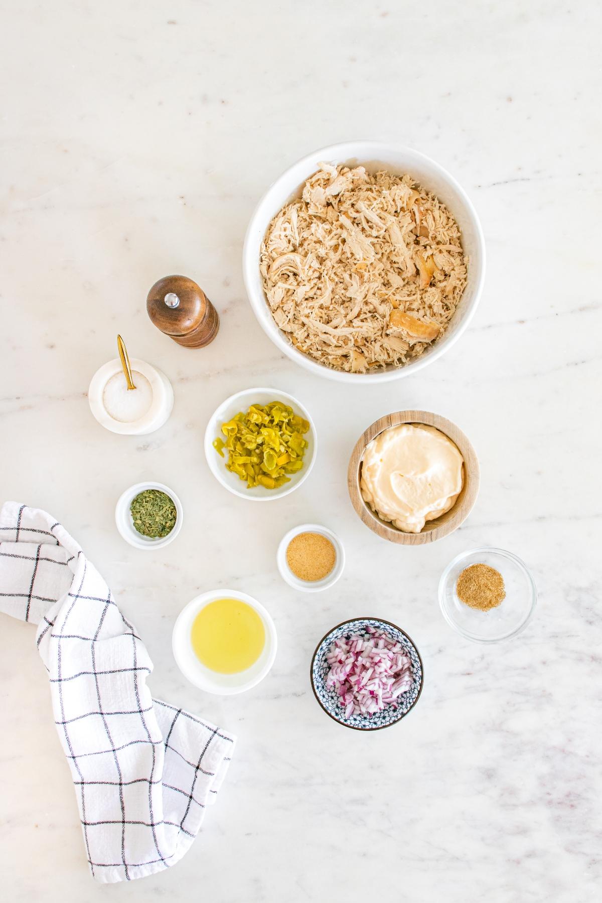 Overhead view of shredded chicken, seasonings, mayo, relish, onions, and a towel on a marble surface.