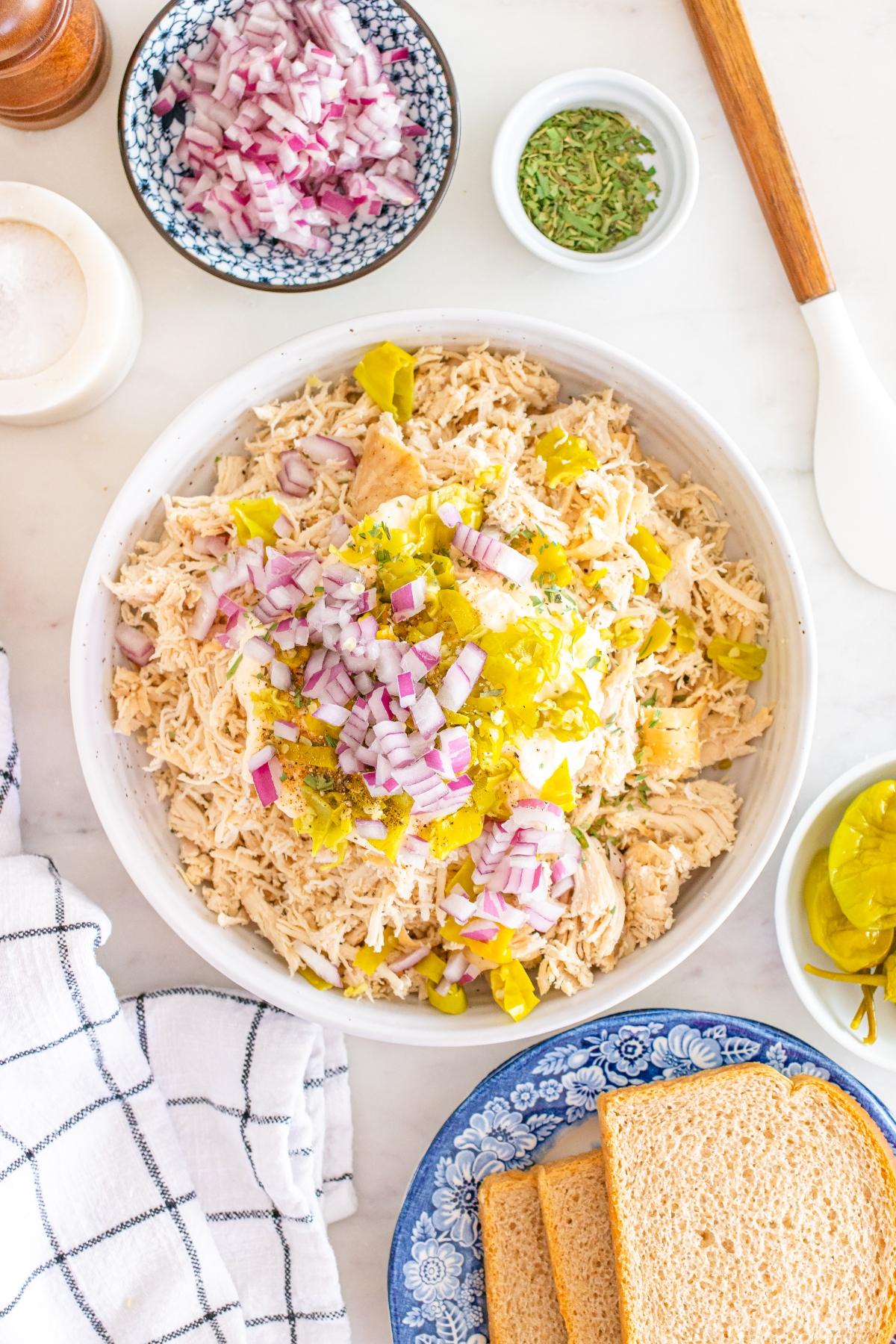 A bowl of shredded chicken topped with diced red onion and pickles, surrounded by bread and seasonings.
