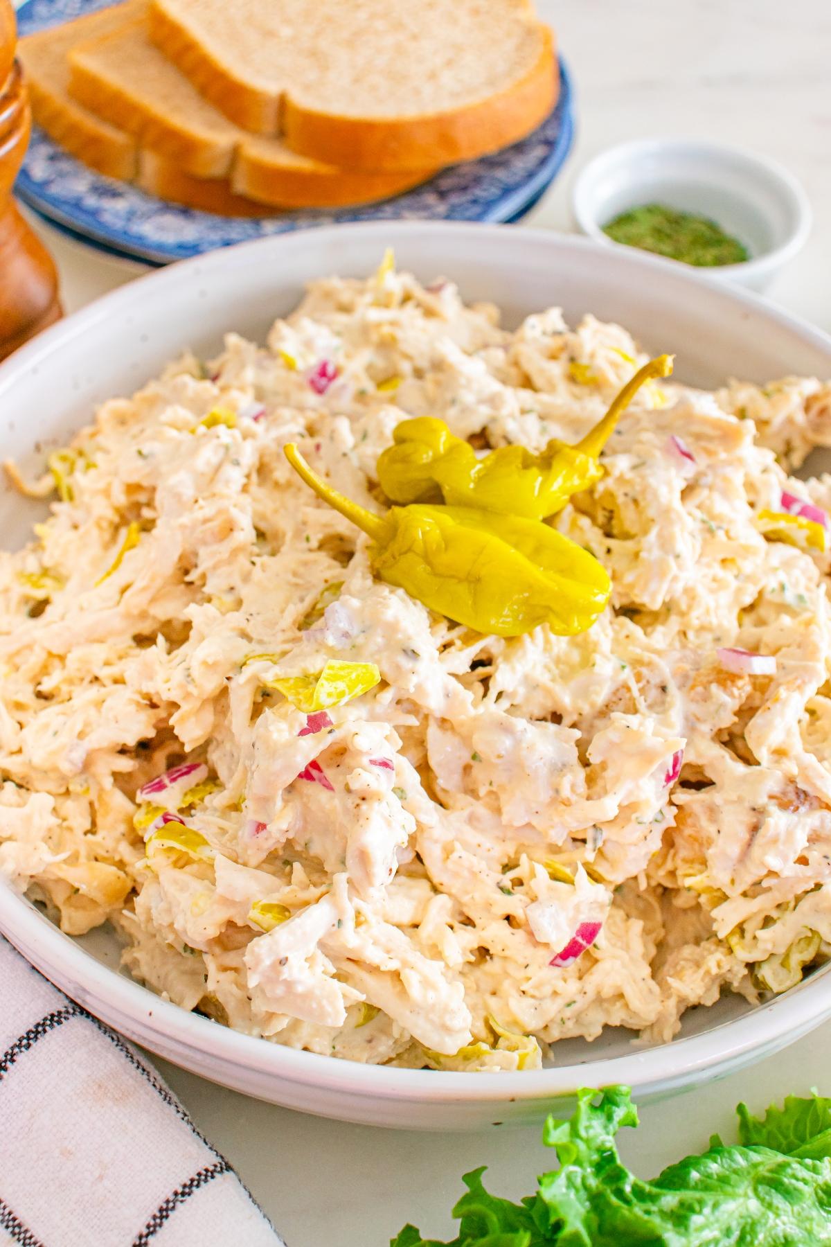 A bowl of creamy Pepperoncini chicken salad topped with pepperoncini, with bread slices and lettuce in the background.