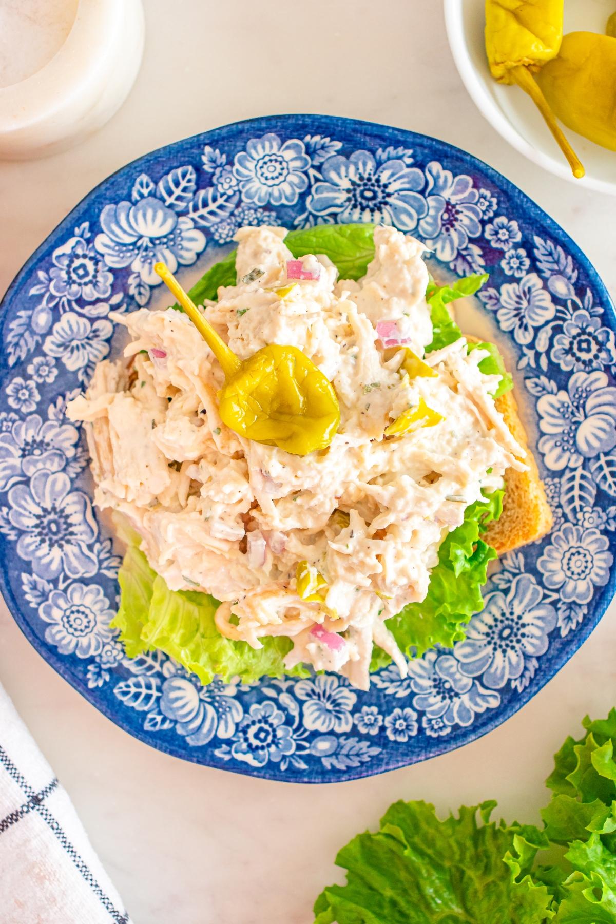 Pepperoncini Chicken salad with chopped onions on lettuce and toast, garnished with a yellow pepper, served on a blue floral plate.