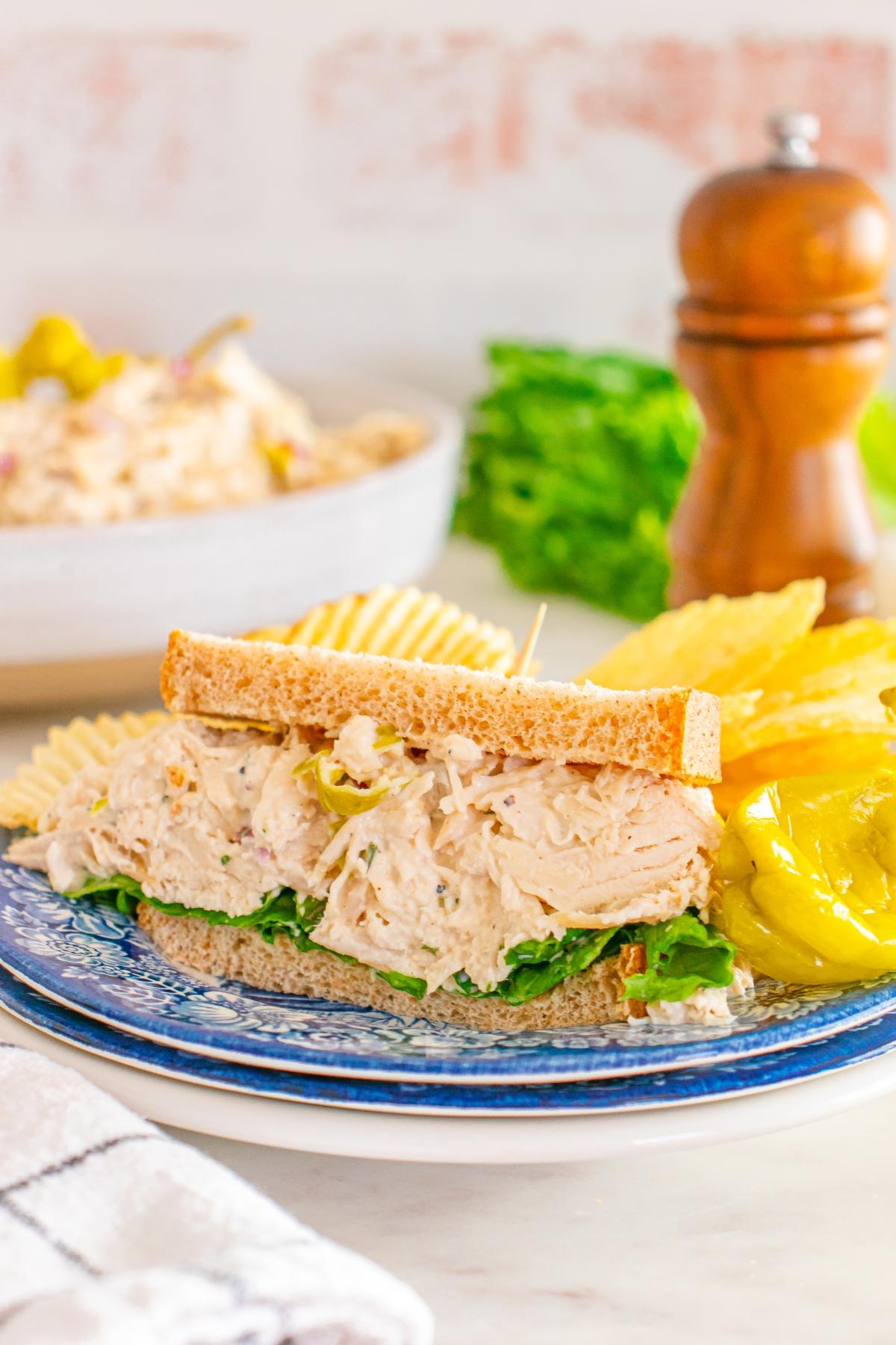 Pepperoncini chicken salad sandwich on wheat bread with lettuce, served with potato chips and a pepper on a blue plate.