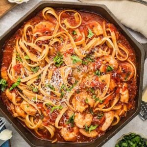 A skillet of Shrimp fra Diavolo linguine in a spicy tomato sauce, topped with parsley and parmesan, with bread and seasonings nearby.