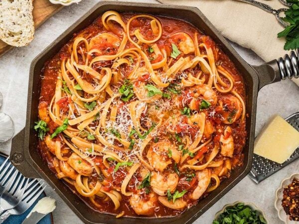 A skillet of Shrimp fra Diavolo linguine in a spicy tomato sauce, topped with parsley and parmesan, with bread and seasonings nearby.