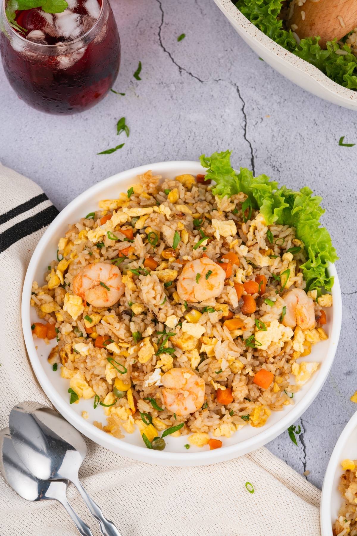 A plate of shrimp fried rice with vegetables, garnished with lettuce, beside a glass of dark drink and utensils.