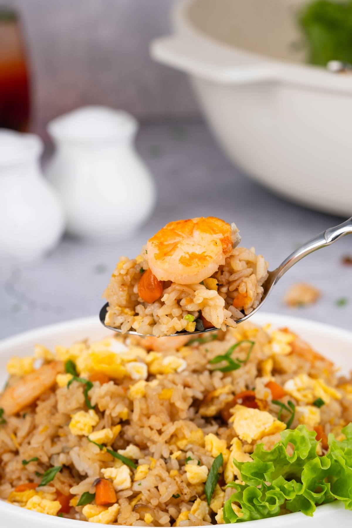 A spoonful of shrimp fried rice with egg and vegetables held above a plate, garnished with lettuce.