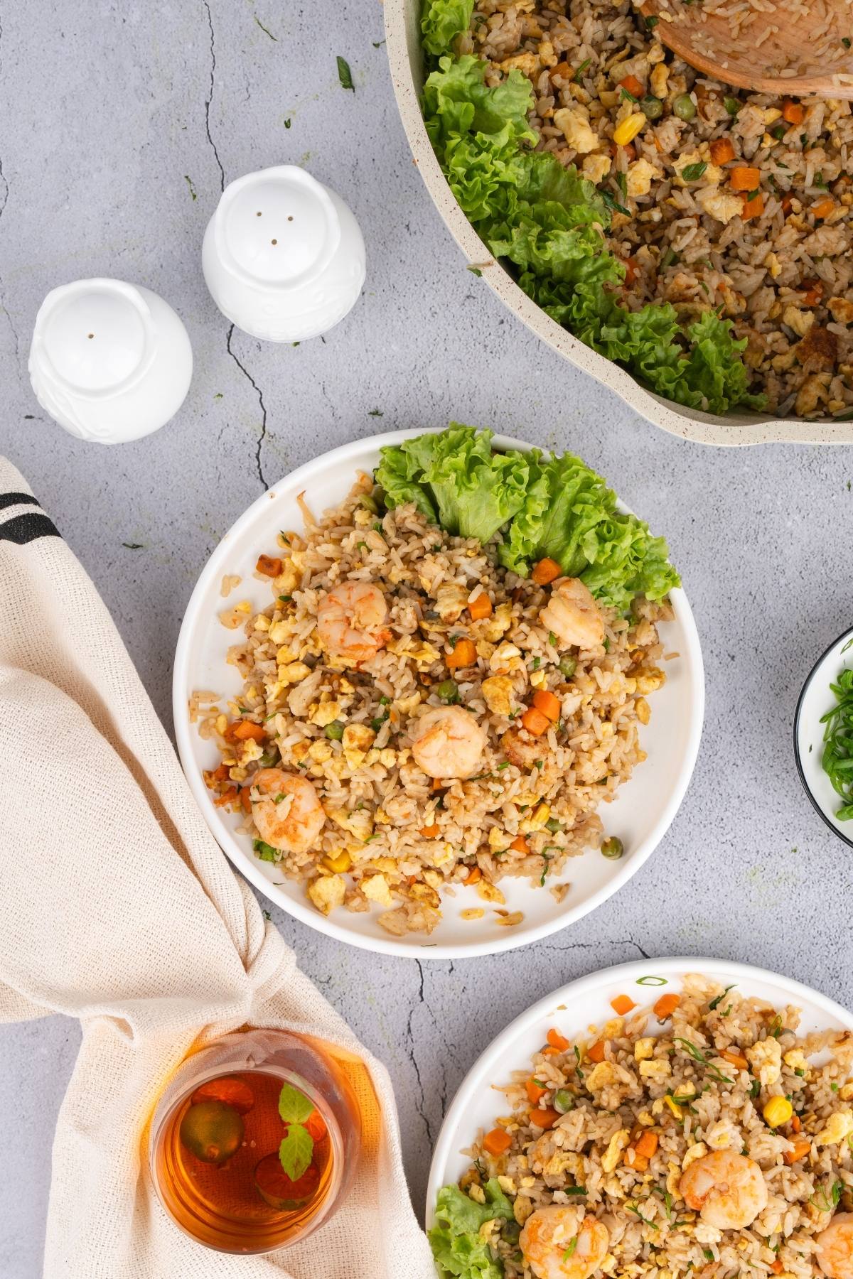 A plate of shrimp fried rice with lettuce, surrounded by a pan of rice, salt and pepper shakers, and a drink.