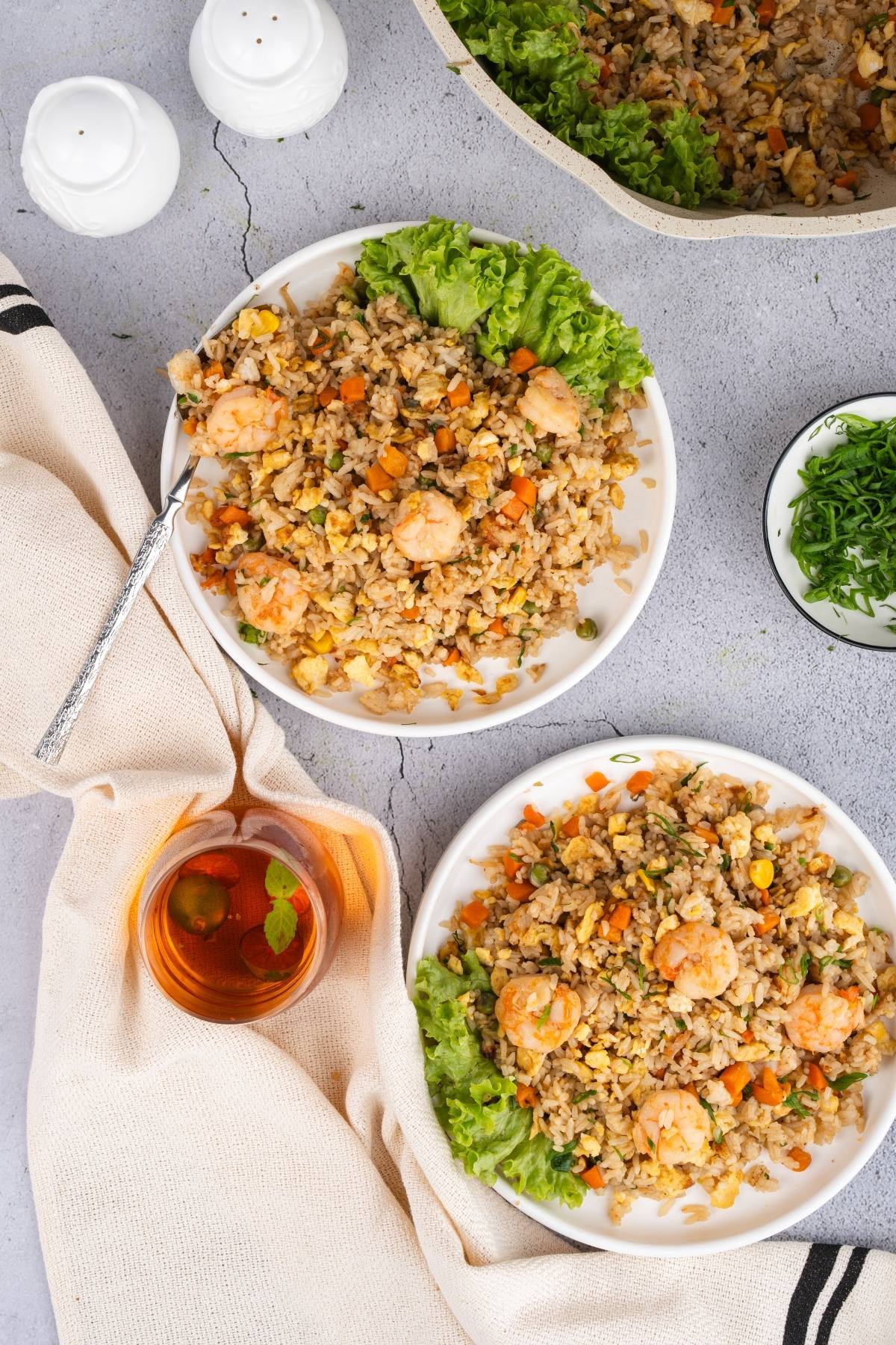 Two plates of shrimp fried rice with vegetables, garnished with lettuce, beside tea and seasonings on a table.