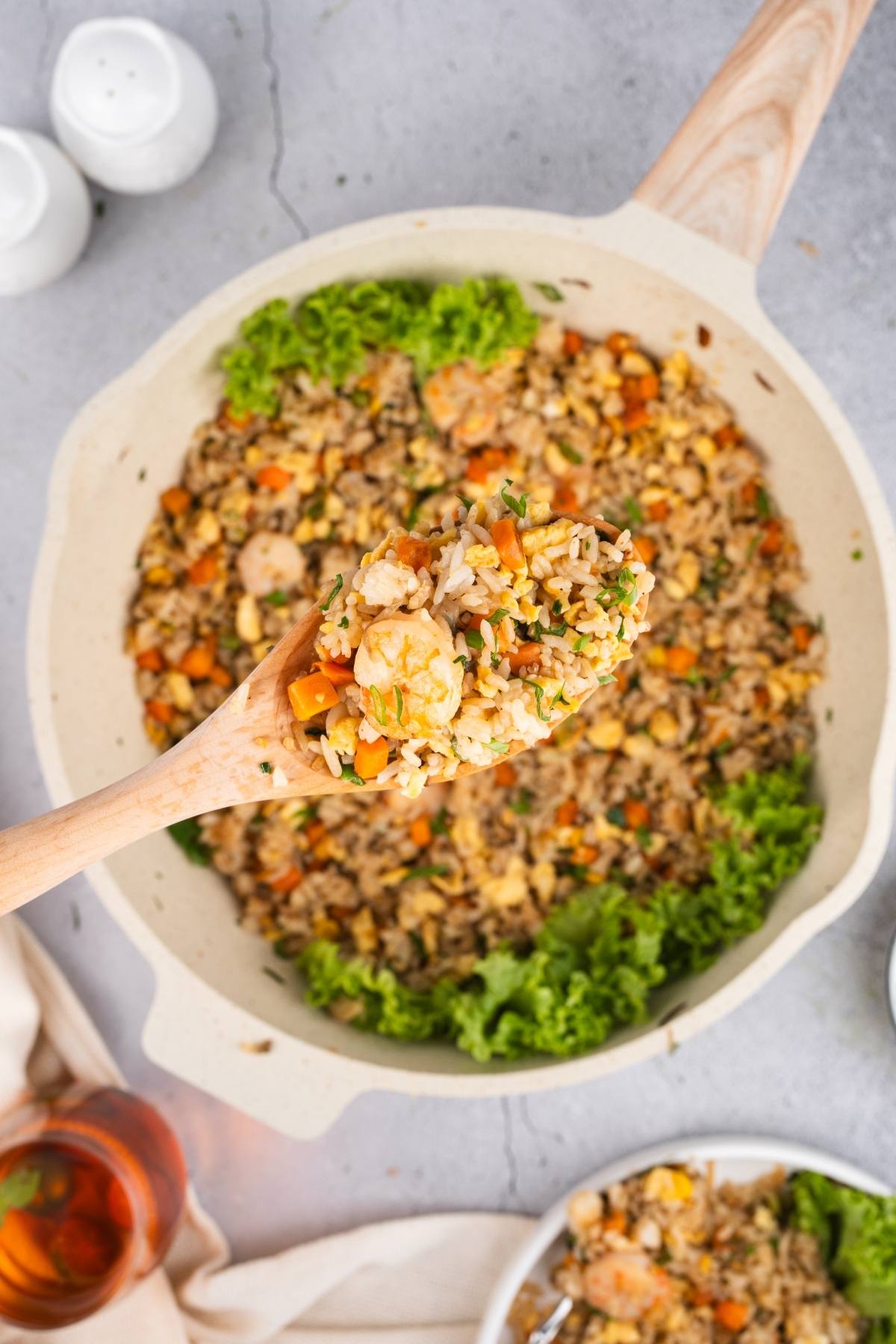 A wooden spoon scoops shrimp fried rice with vegetables from a large pan, garnished with leafy greens.