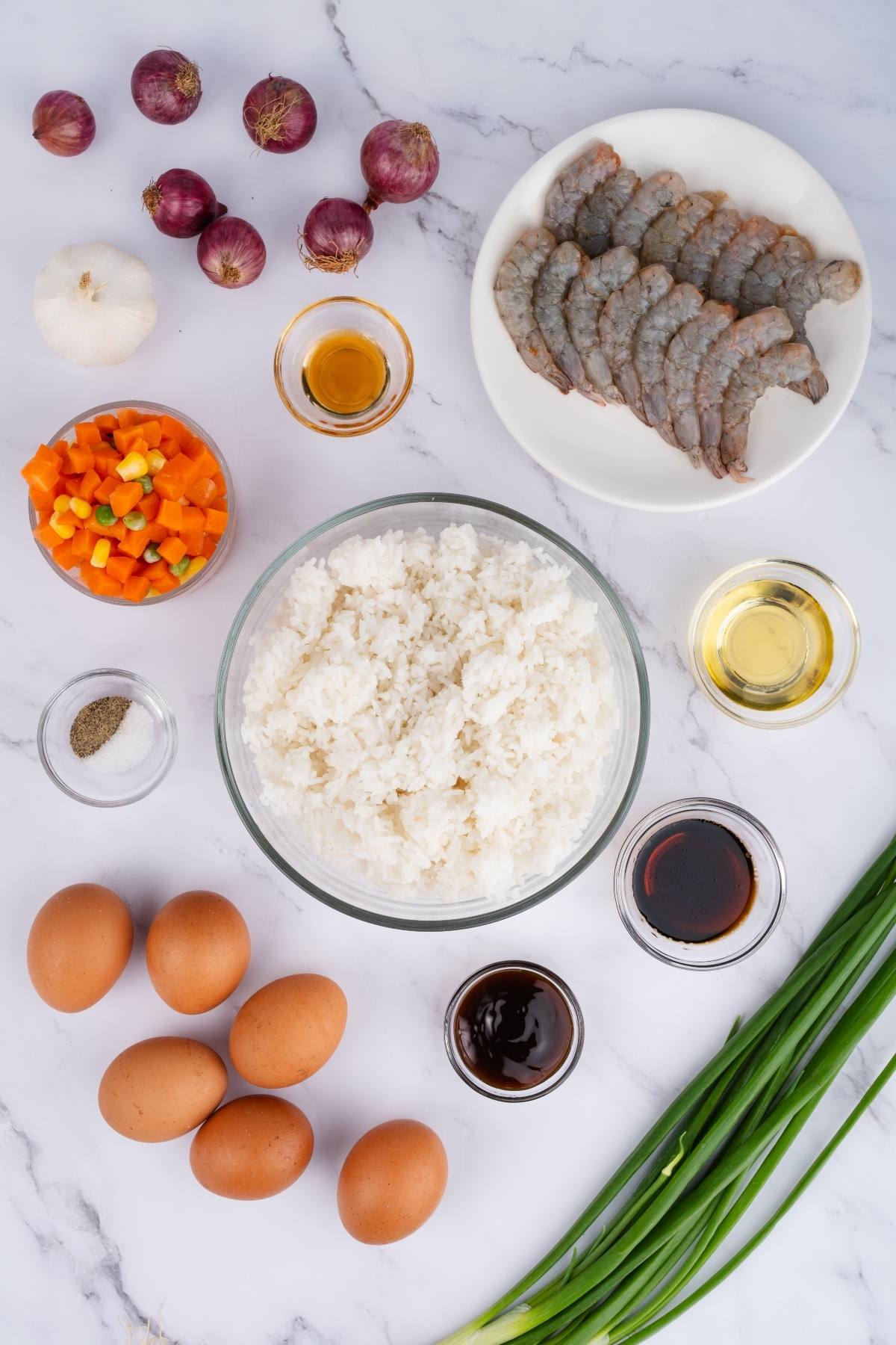 Top view of shrimp, eggs, rice, chopped vegetables, sauces, onions, garlic, and green onions on a white surface.