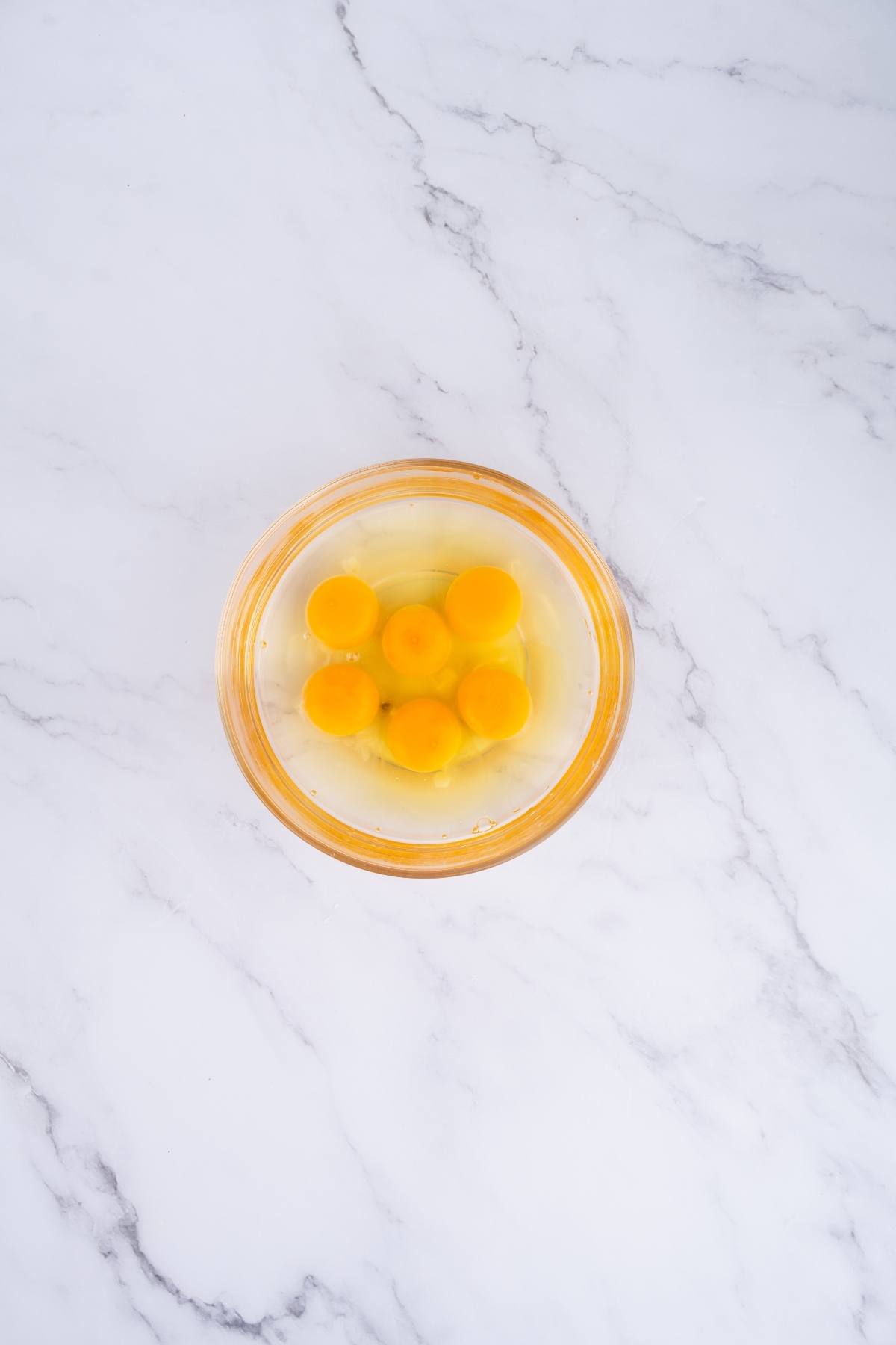 A glass bowl with six cracked eggs on a white marble surface, viewed from above.