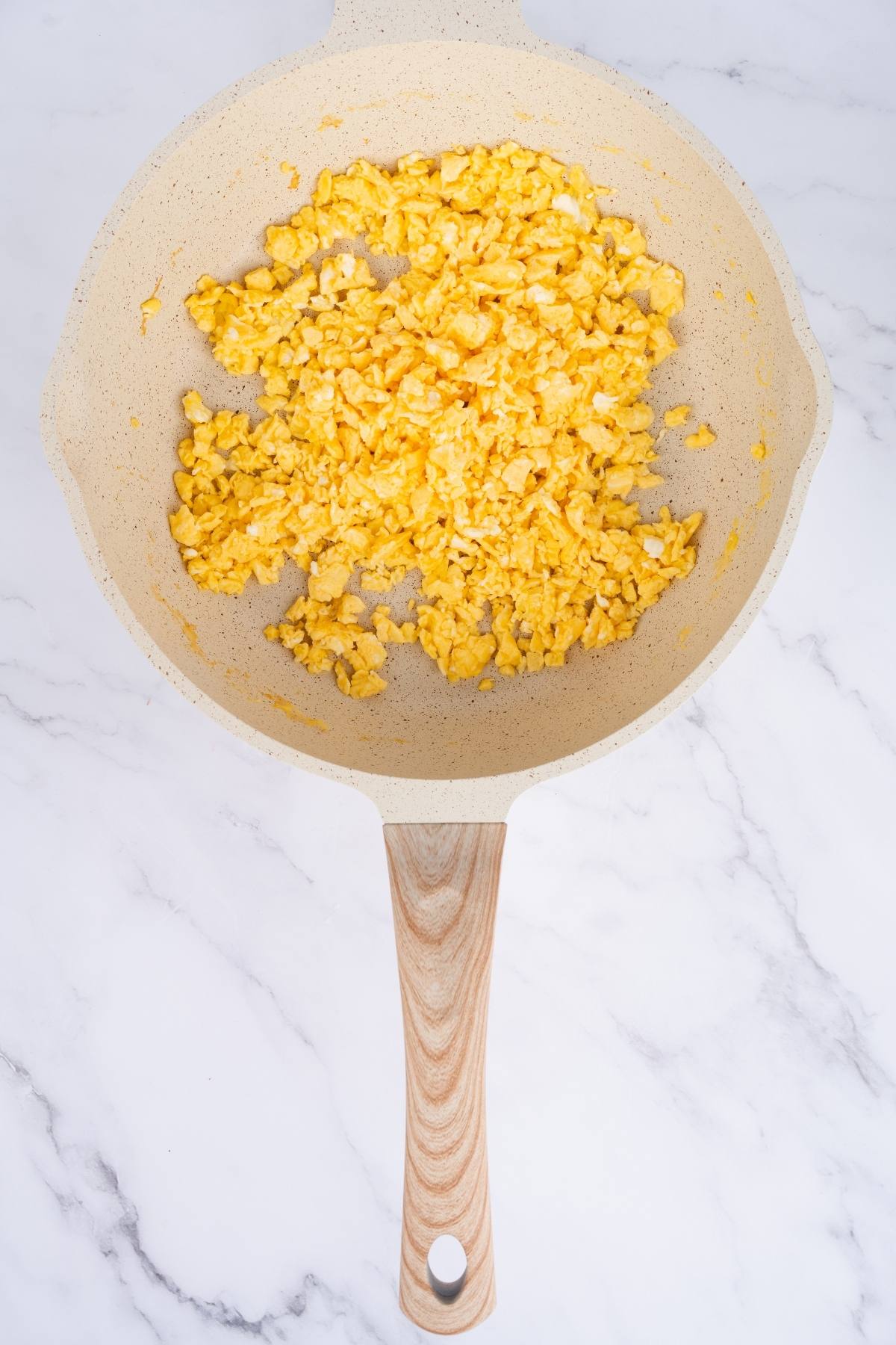 Scrambled eggs in a beige non-stick frying pan with a wooden handle on a marble countertop.