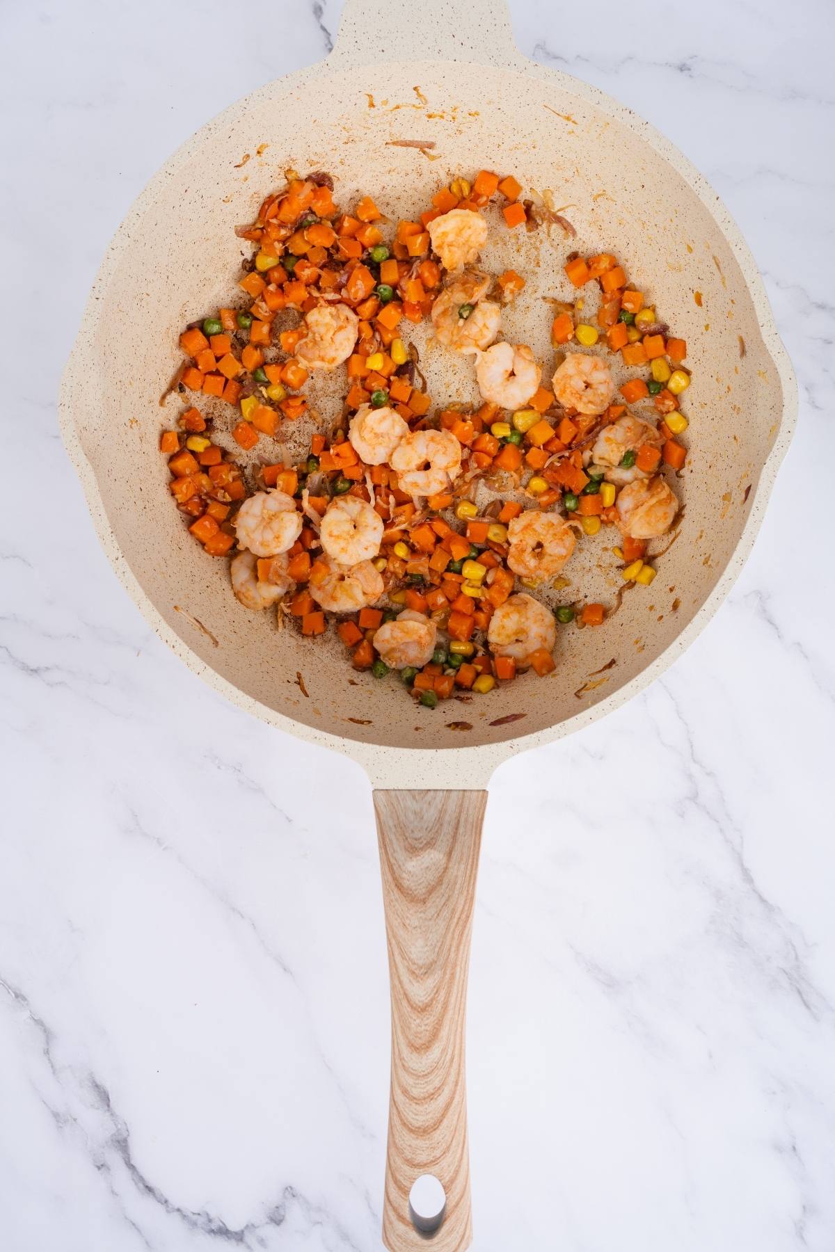 Shrimp, diced carrots, corn, and peas cooking in a beige skillet on a marble countertop.