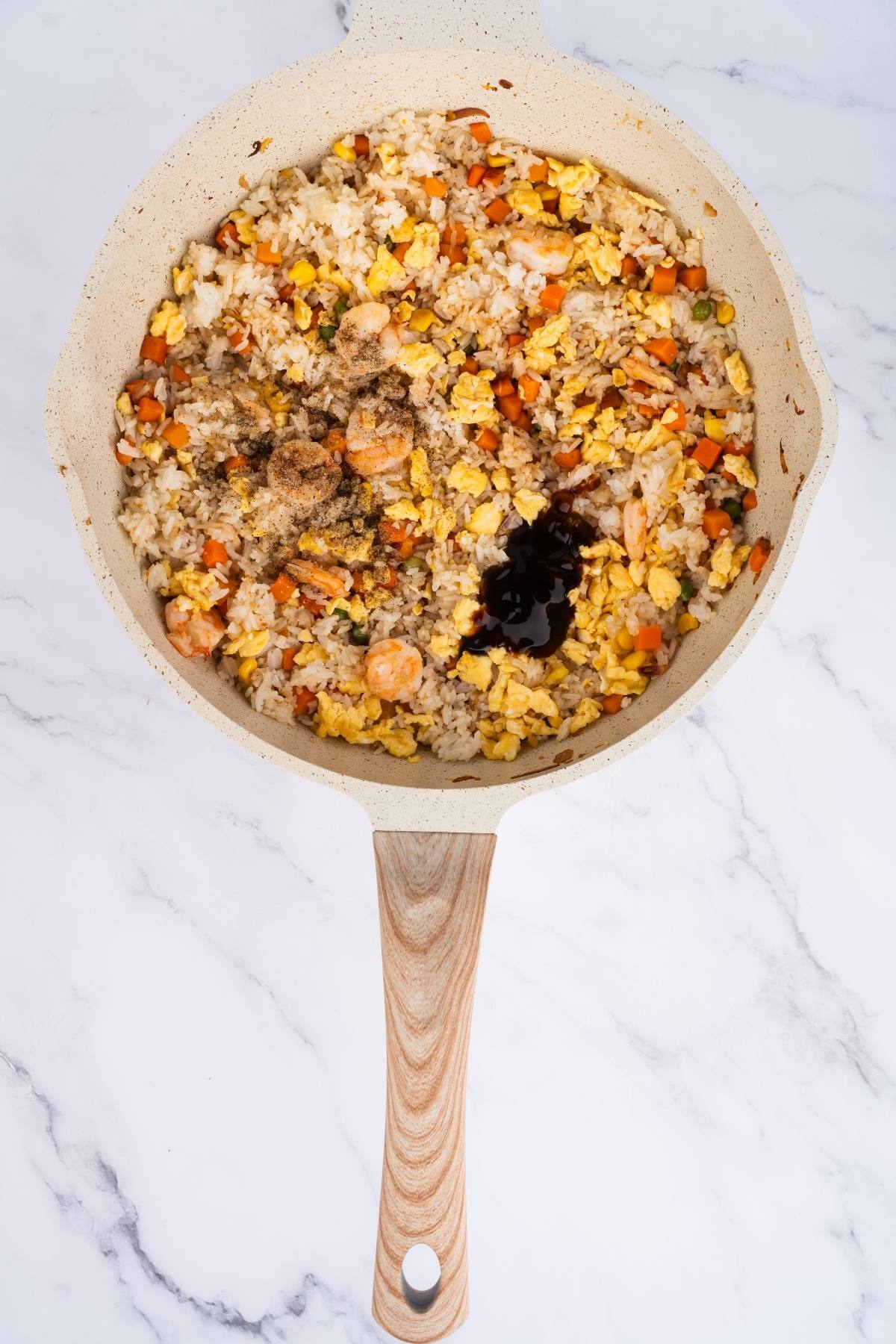 A skillet with fried rice, shrimp, scrambled eggs, vegetables, and soy sauce on a marble surface.