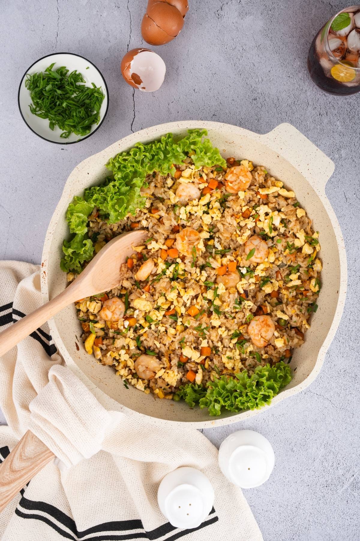 A pan of shrimp fried rice with eggs, carrot, lettuce, and a wooden spoon on a gray tabletop.