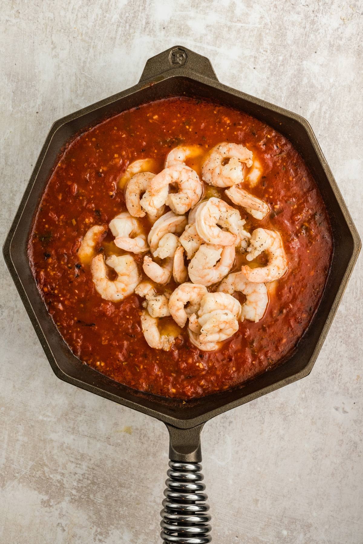 Shrimp cooking in a skillet filled with tomato-based sauce, seen from above on a light surface.