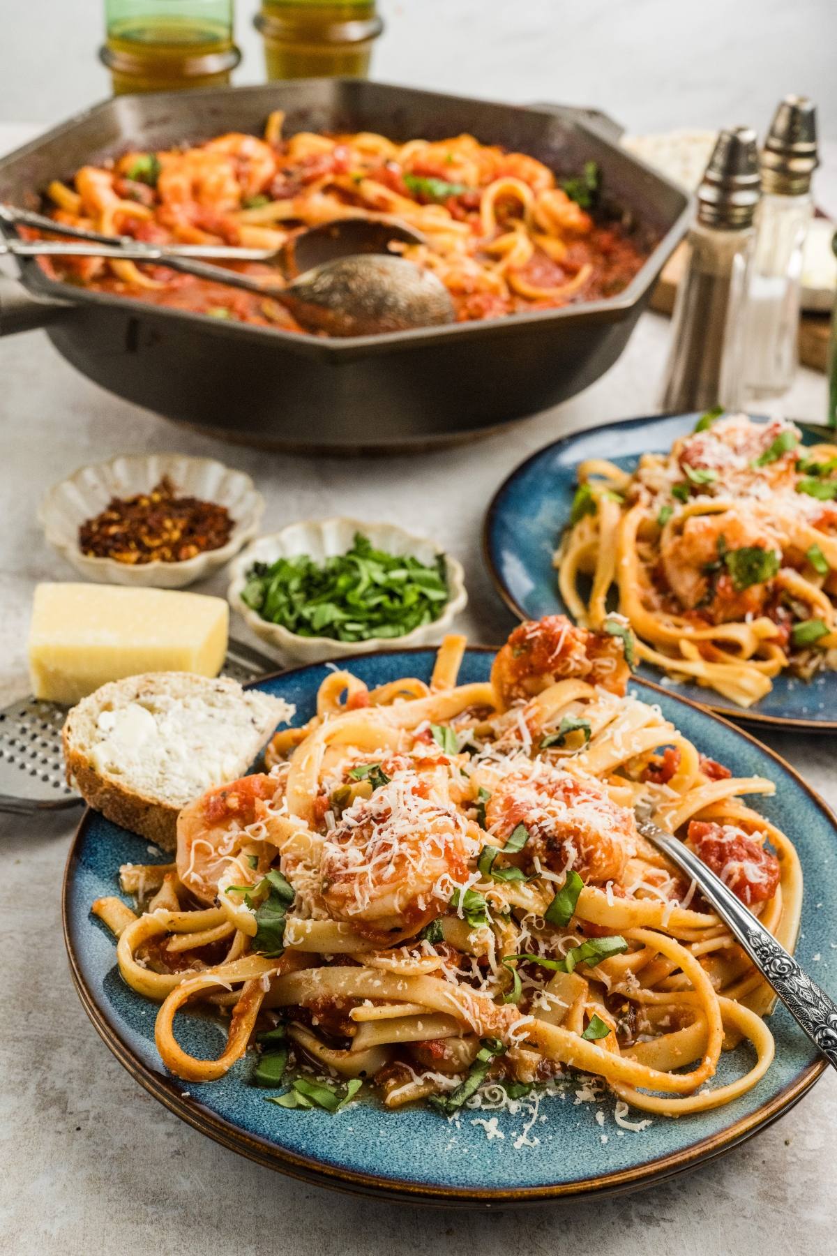 Two plates of Shrimp fra diavolo pasta with tomato sauce and cheese, bread, and fresh herbs, with a skillet of pasta in the background.