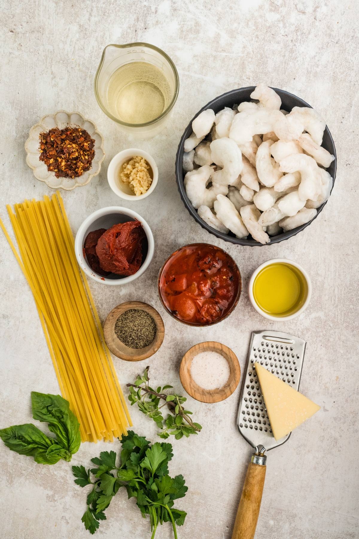 Uncooked shrimp, pasta, tomato sauce, oil, cheese, herbs, and spices arranged on a white surface.