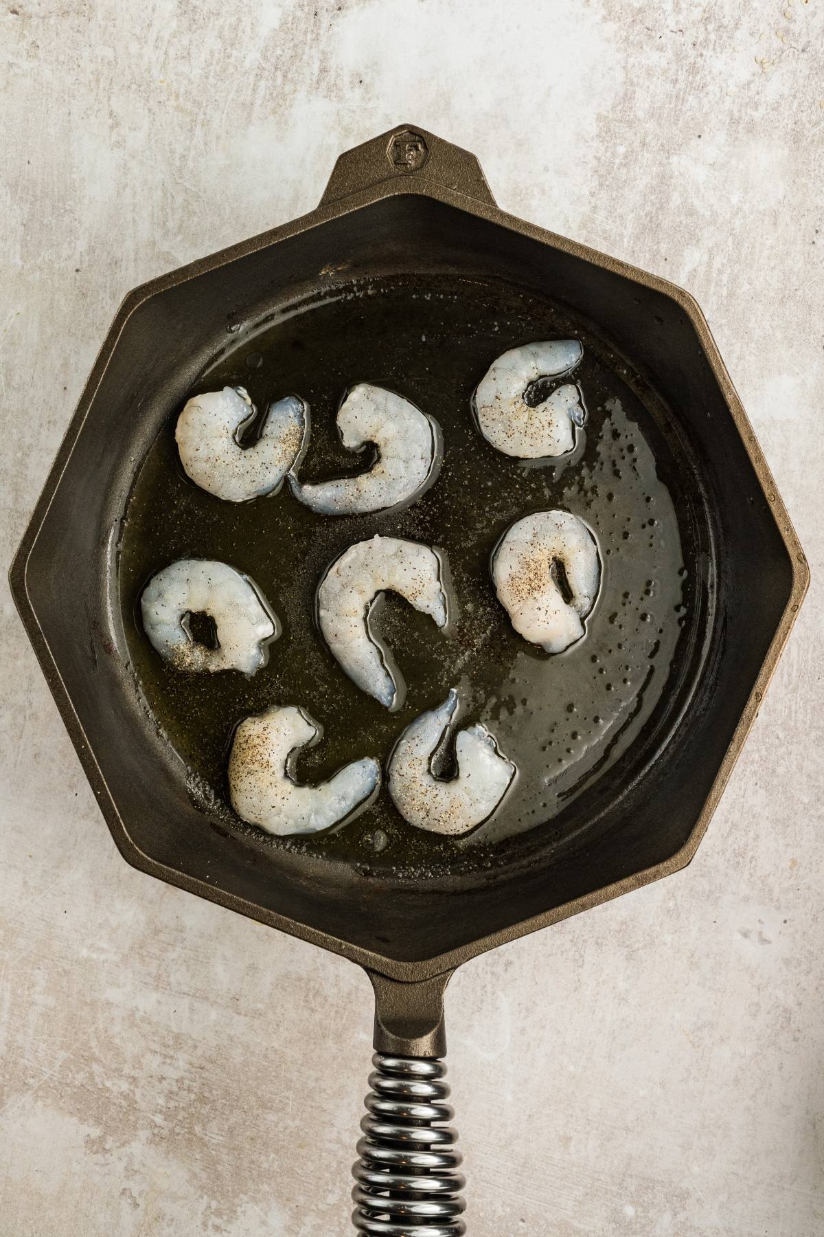 Raw shrimp cooking in oil inside a hexagonal cast iron skillet on a light-colored surface.