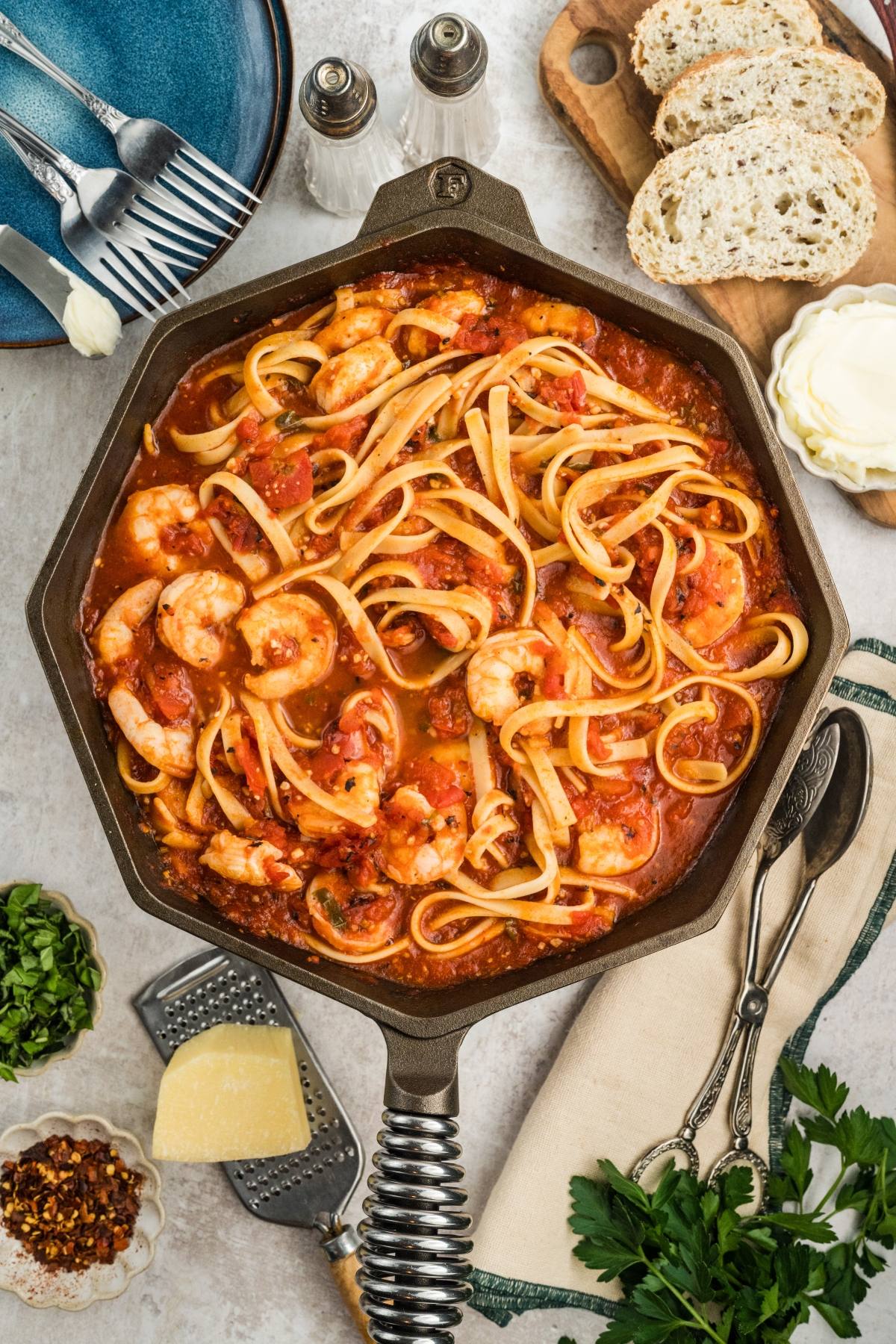 A skillet of shrimp pasta in tomato sauce, surrounded by bread, herbs, cheese, and utensils on a table.