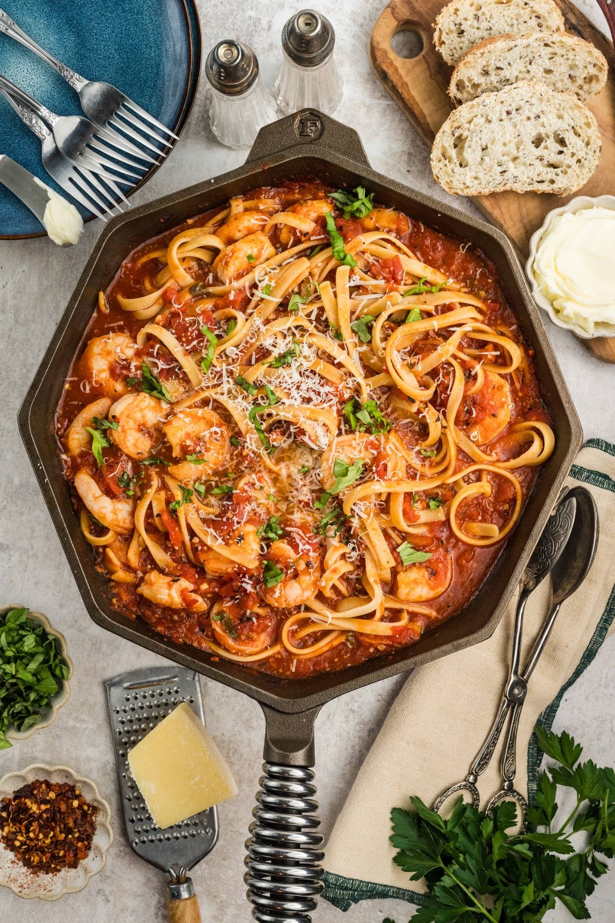 Skillet of Shrimp fra diavolo pasta in tomato sauce, topped with parsley and cheese, with bread and utensils on the side.